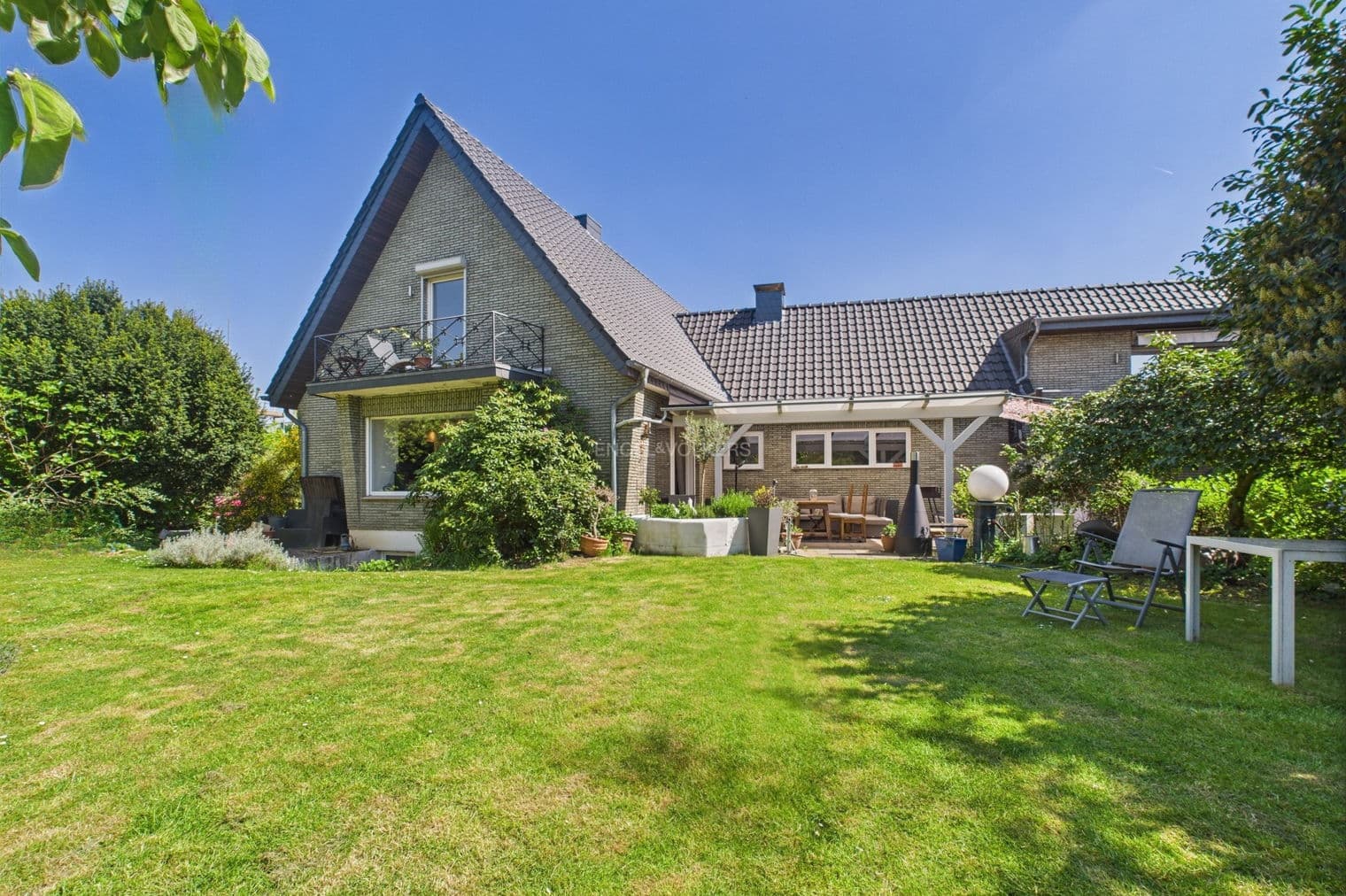 Exterior view of a two-story house with a green lawn, trees, and a blue sky. The house has a gray brick facade and a dark gray roof.