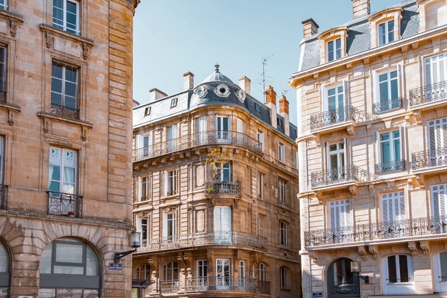 Europäische Stadtstraße mit hellbraunen Steingebäuden, Fenstern und Balkonen unter einem klaren blauen Himmel.