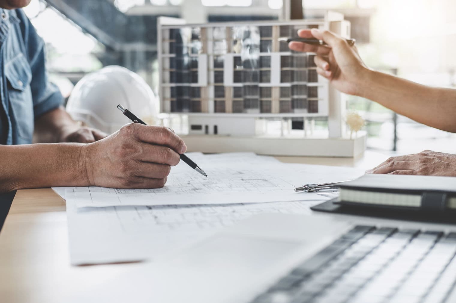 Architekten überprüfen Baupläne und ein Gebäudemodell und verwenden Stifte, um Details auf den Plänen zu markieren. Ein weißer Schutzhelm liegt in der Nähe.