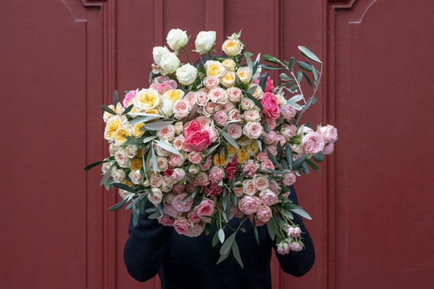 Person in black coat holding a large bouquet of pink, white, and yellow roses with olive branches against a red door.