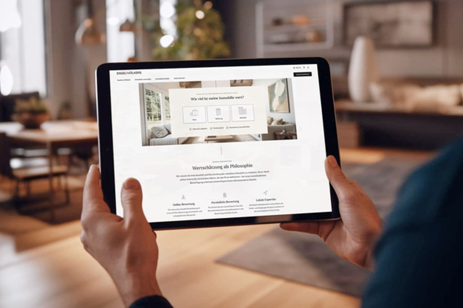 Ein Mann hält ein Tablet, auf dem die Engel & Völkers Immobilien-Website in einem modernen Wohnzimmer angezeigt wird.