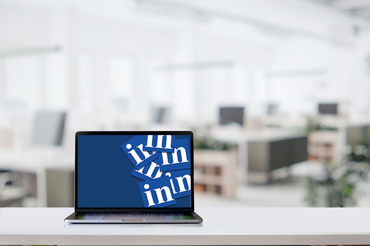 Laptop on a white desk displays the LinkedIn logo against a blurred office background.