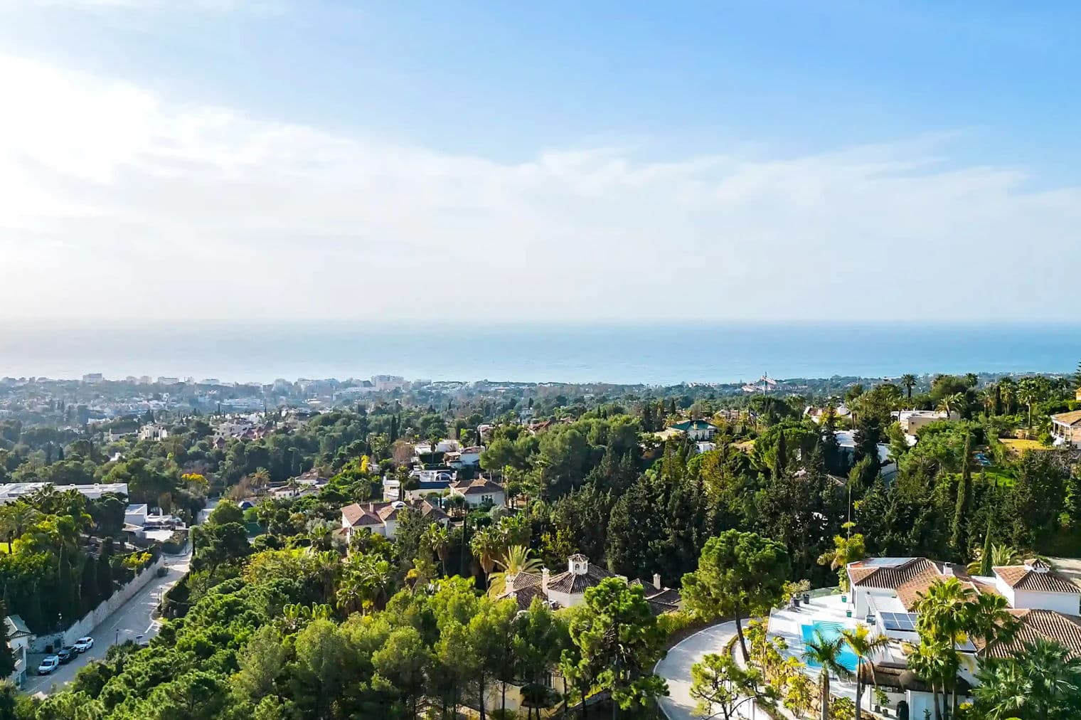 Vista panoràmica d'un poble costaner amb arbres de color verd exuberant, cases blanques i un oceà blau sota un cel clar.