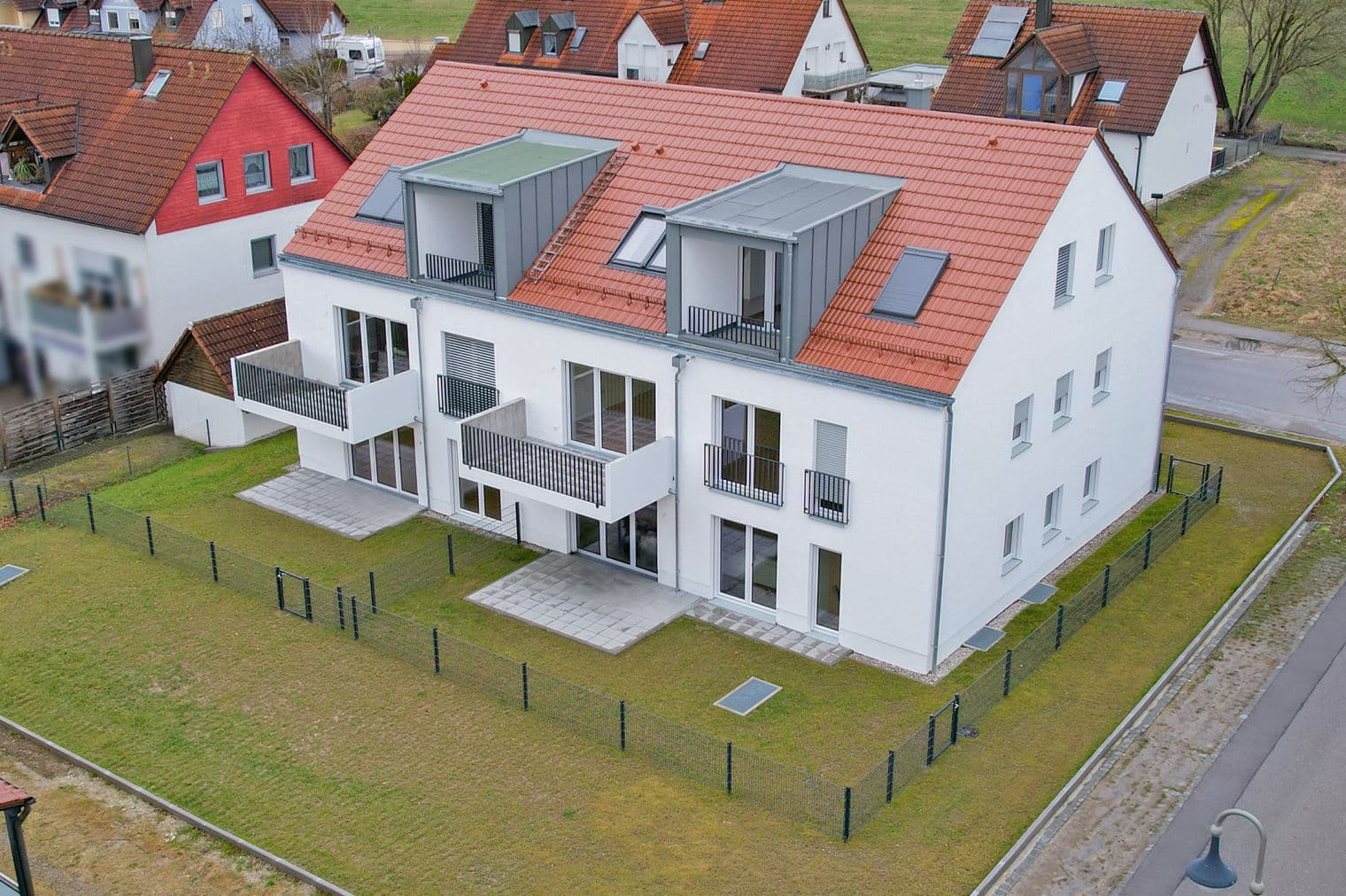 Aerial view of a modern, white apartment building with a red tile roof and gray balconies, surrounded by a green lawn and a fence.