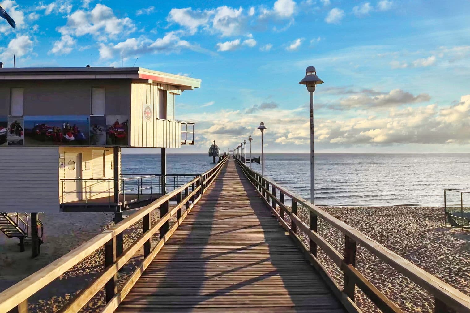 Hölzerner Pier, der sich unter blauem Himmel mit Wolken ins Meer erstreckt. Eine Rettungsschwimmerstation steht am Strand.