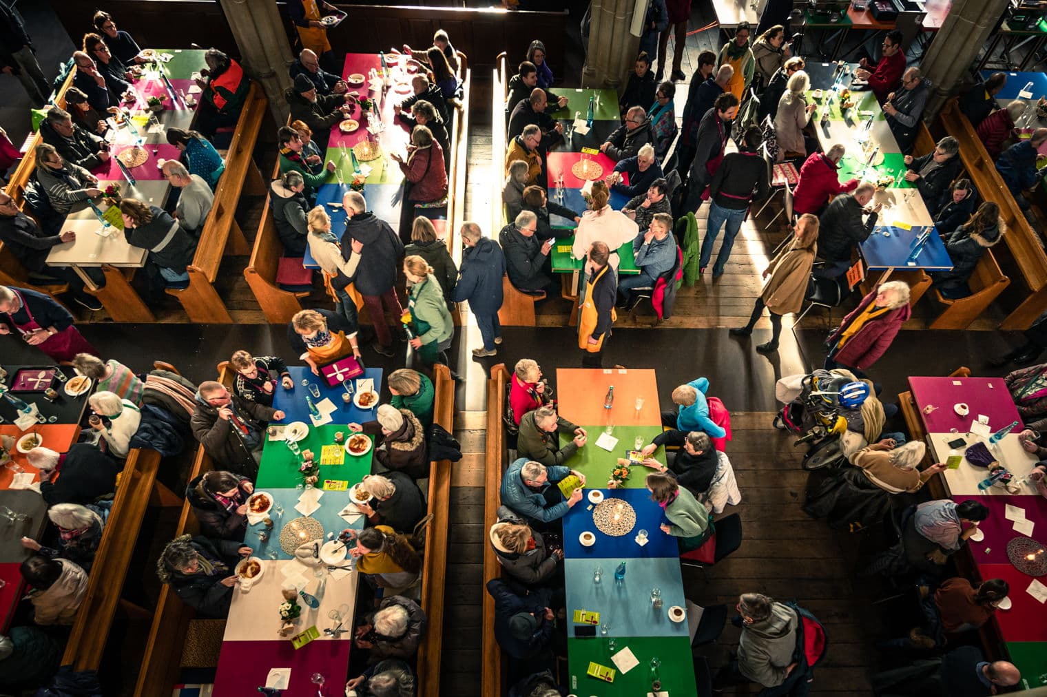 Luftaufnahme eines überfüllten Raumes mit Menschen, die an bunten Tischen sitzen, essen und sich unterhalten. Holzbänke säumen die Tische.