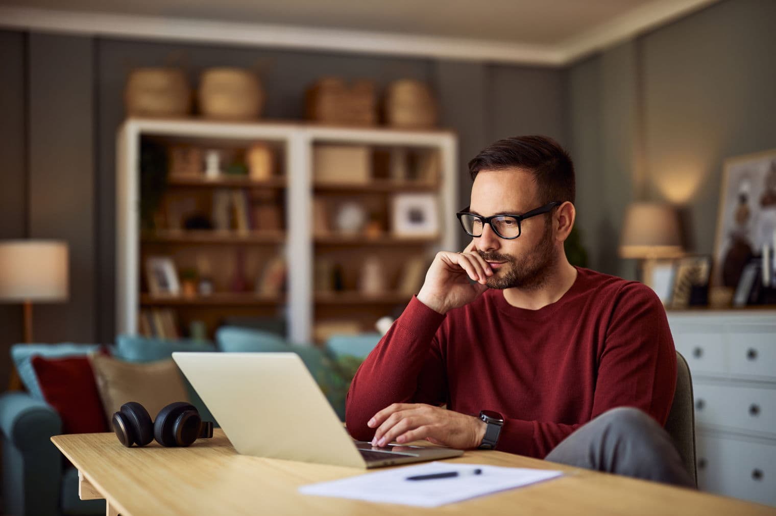 Mann mit rotem Pullover arbeitet an einem Laptop an einem Holzschreibtisch in einem Wohnzimmer.