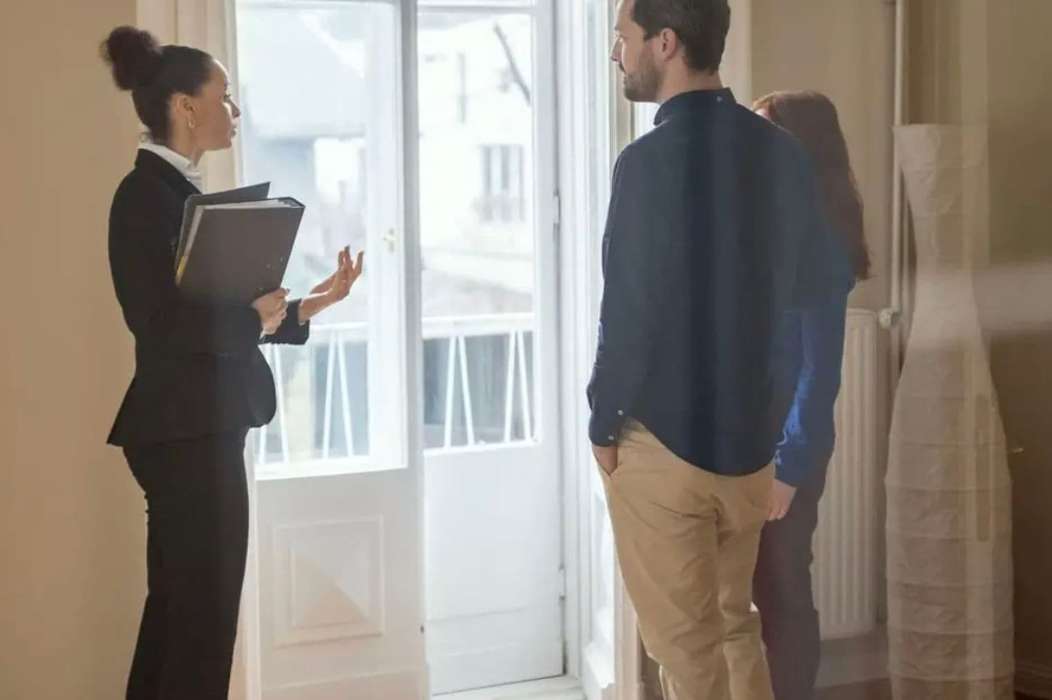 Un agent immobilier en costume noir fait un geste vers un couple dans une pièce lumineuse avec une porte blanche. Elle tient un dossier noir.