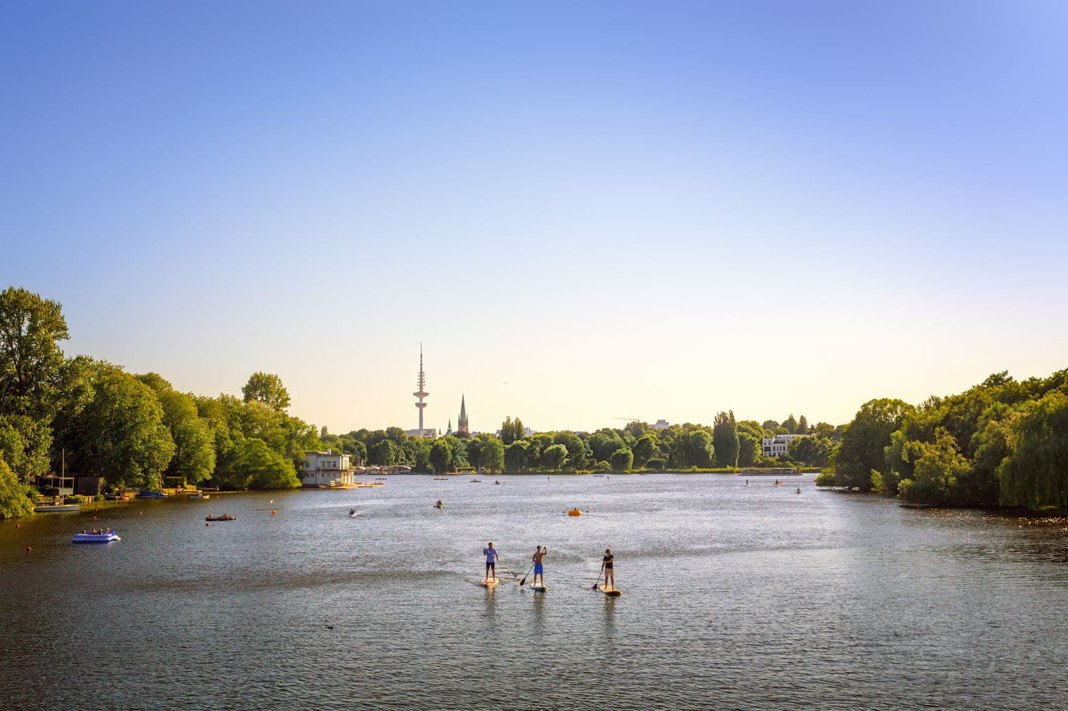 Drei Personen paddeln auf einem Fluss unter einem klaren, blauen Himmel. Grüne Bäume säumen die Ufer, mit einer Stadtsilhouette in der Ferne.