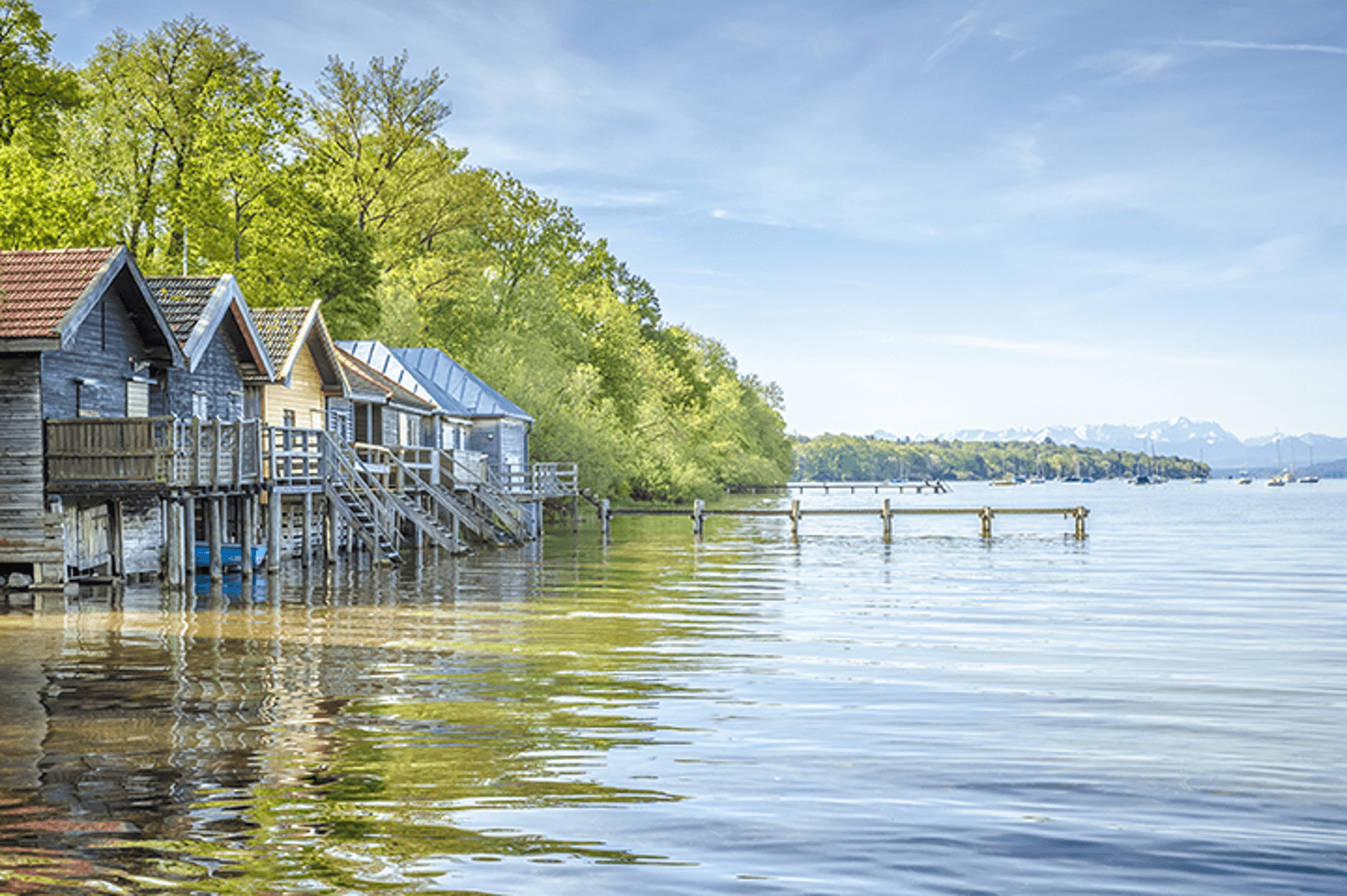 Scenic view of wooden boathouses on a lake, with green trees and distant mountains under a blue sky.