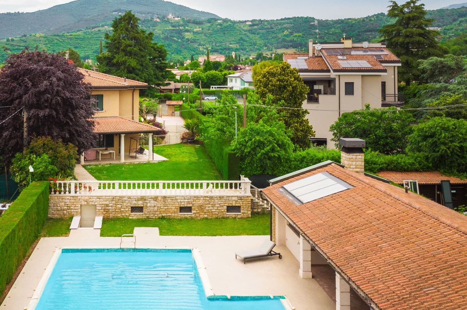 Luftaufnahme eines blauen Schwimmbeckens, Ziegeldächer und grüne Bäume mit Bergen im Hintergrund.