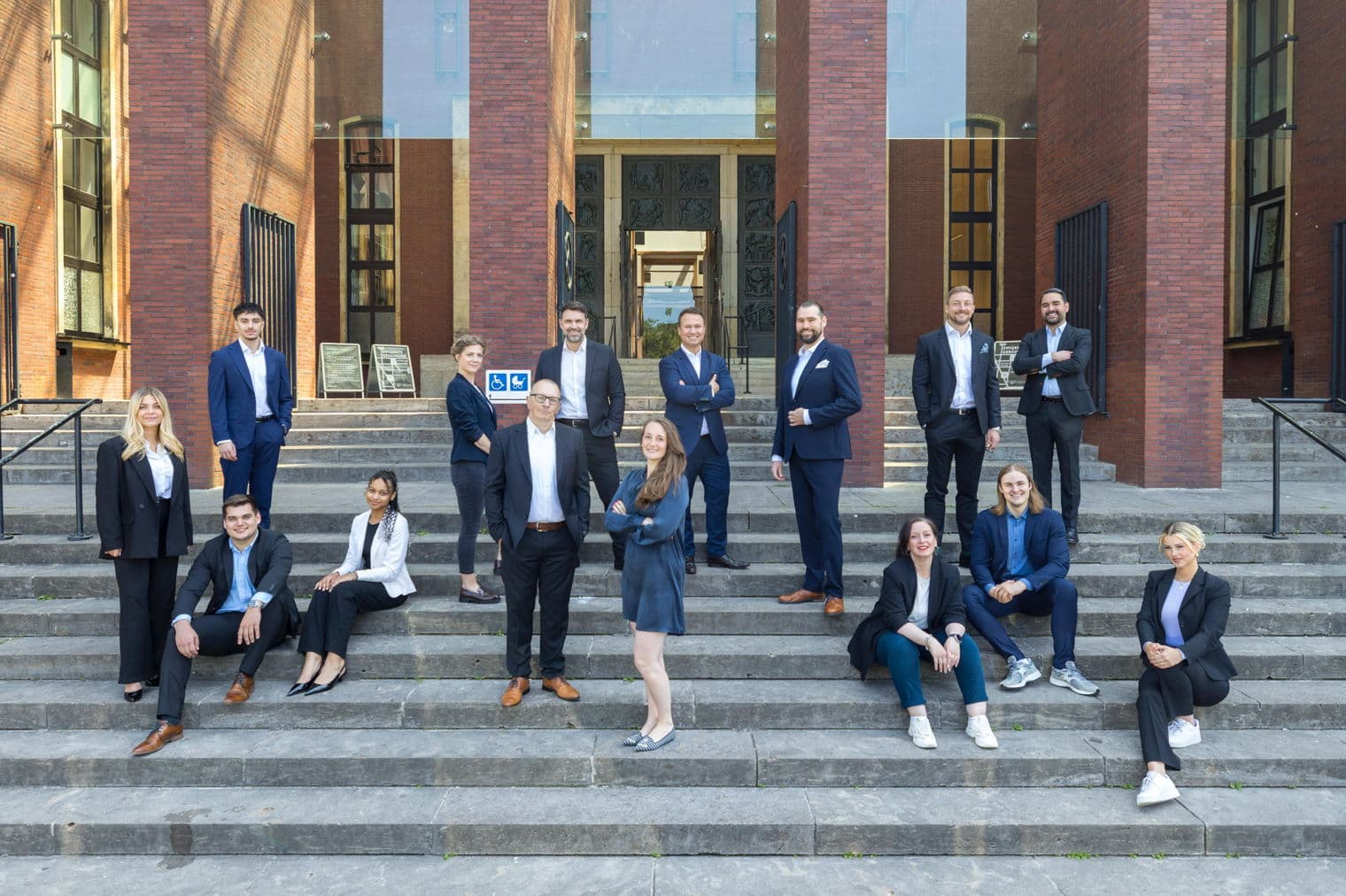 Eine Gruppe von 15 Immobilienmaklern posiert auf Steinstufen vor einem Backsteingebäude mit Glasfenstern und -türen.