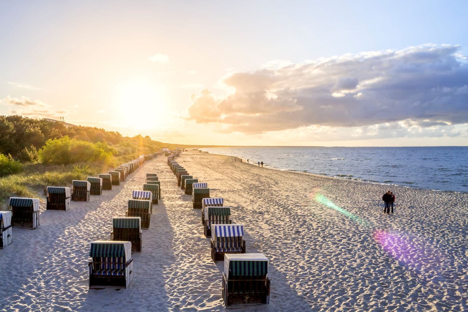 Strandszene mit Reihen von gestreiften Strandstühlen, die unter einem hellen, sonnigen Himmel zum Meer führen. Zwei Personen stehen in der Nähe der Wasserkante.