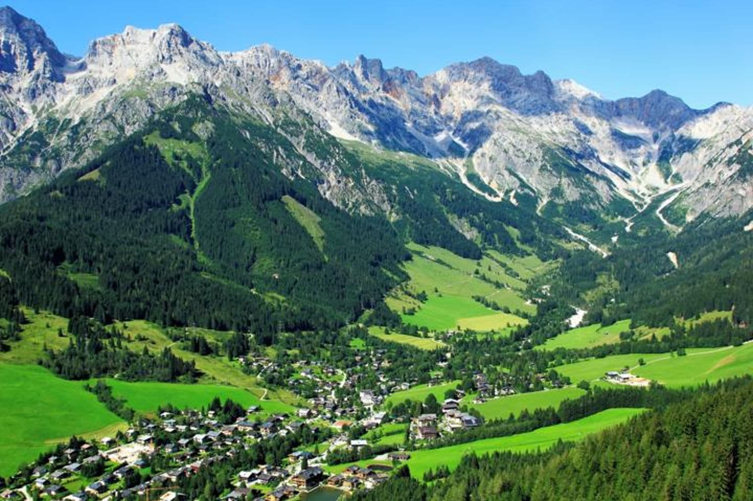 Malerischer Blick auf ein Dorf, eingebettet in ein Tal, umgeben von grünen Wäldern und hoch aufragenden, schneebedeckten Bergen unter einem klaren blauen Himmel.