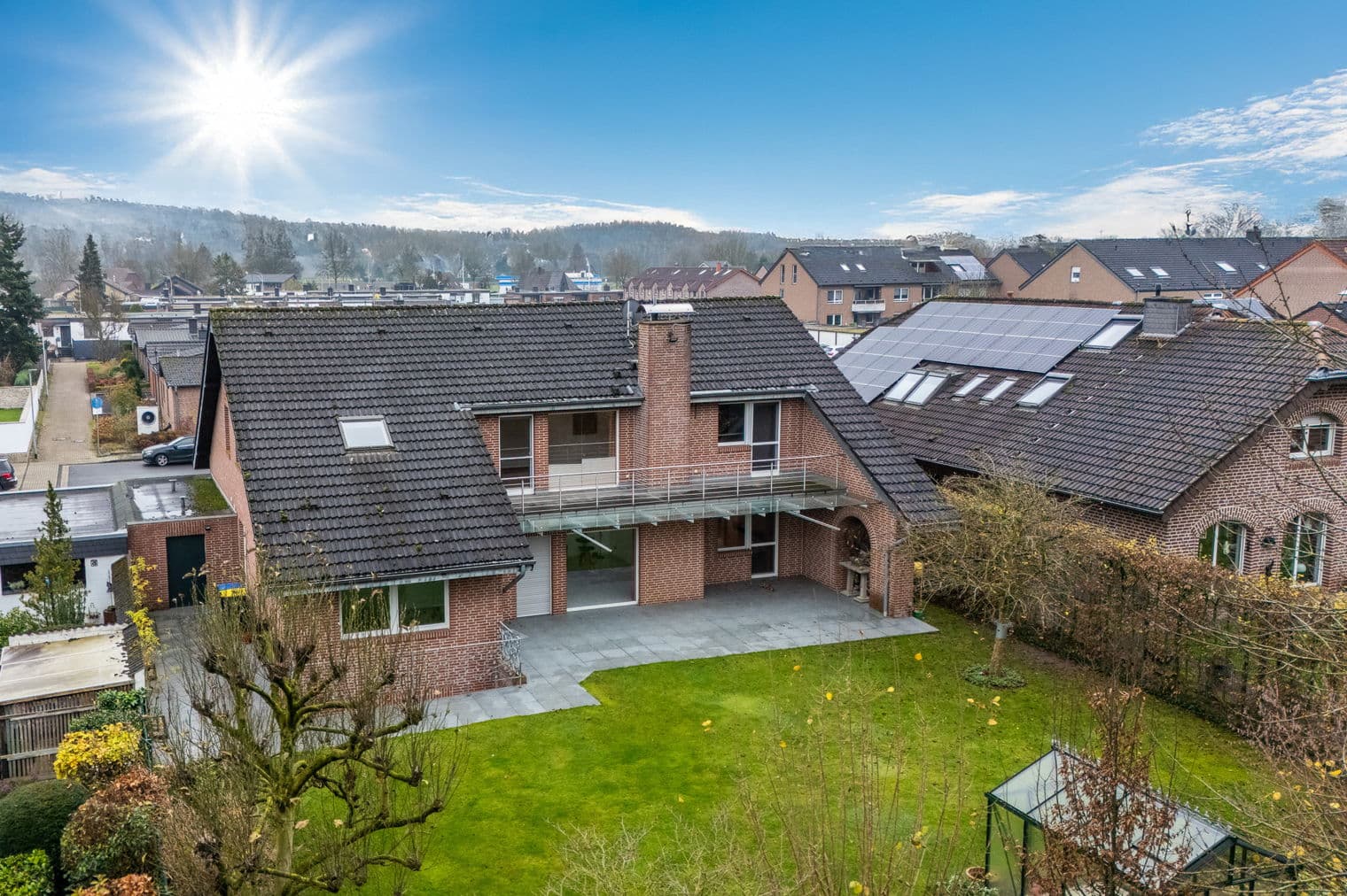 Aerial view of a red brick house with a dark roof, green lawn, patio, and greenhouse under a bright blue sky.