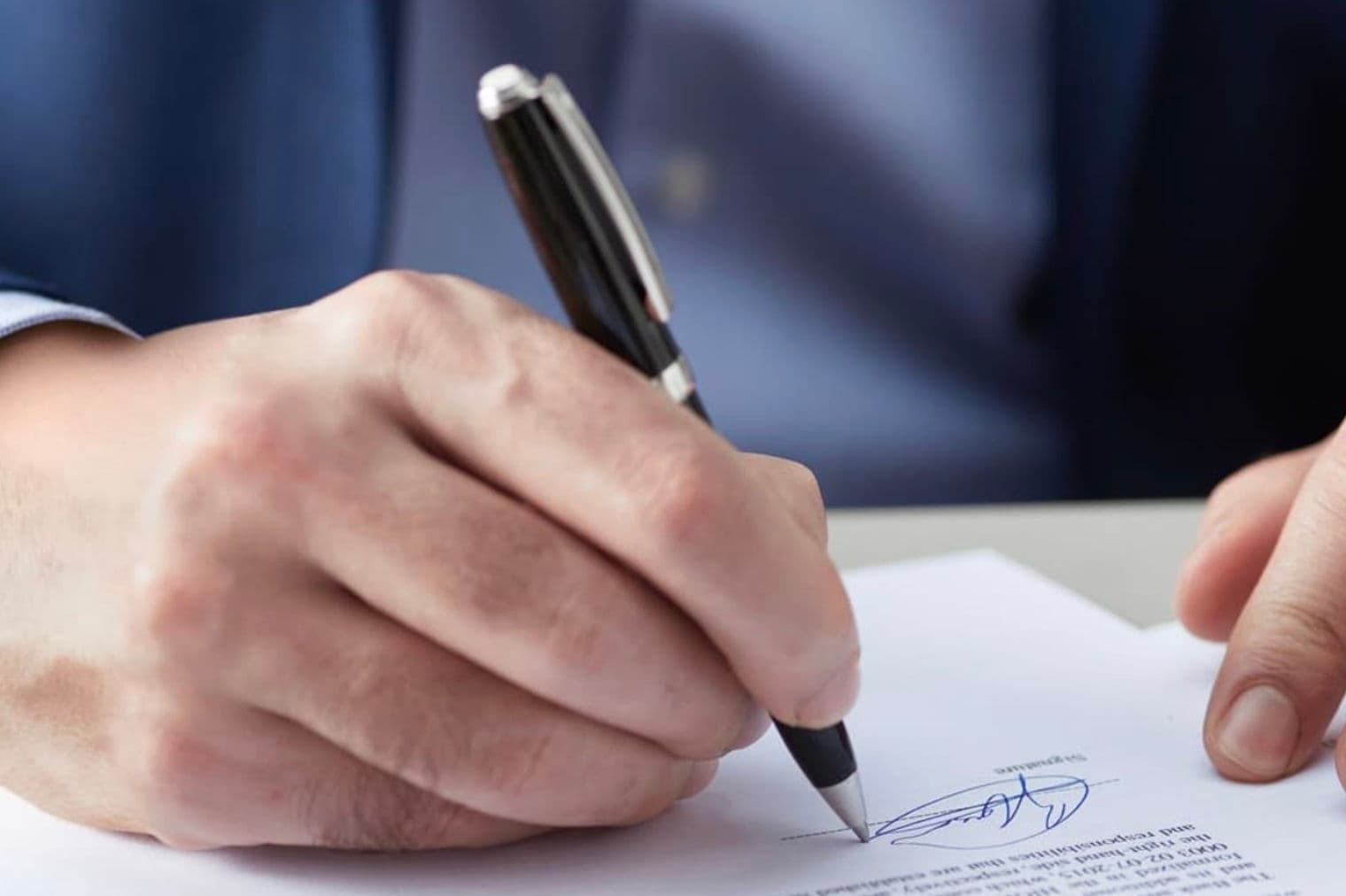Une personne en costume bleu signe un document avec un stylo noir.