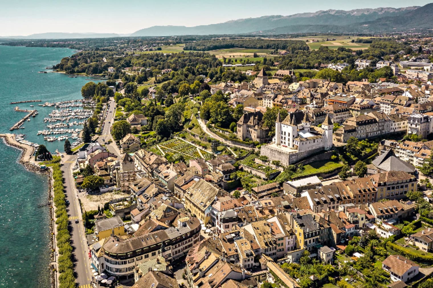 Vista aerea di Nyon, Svizzera, che mostra un porto turistico con barche, un castello bianco ed edifici con tetti di tegole rosse. Montagne sullo sfondo.