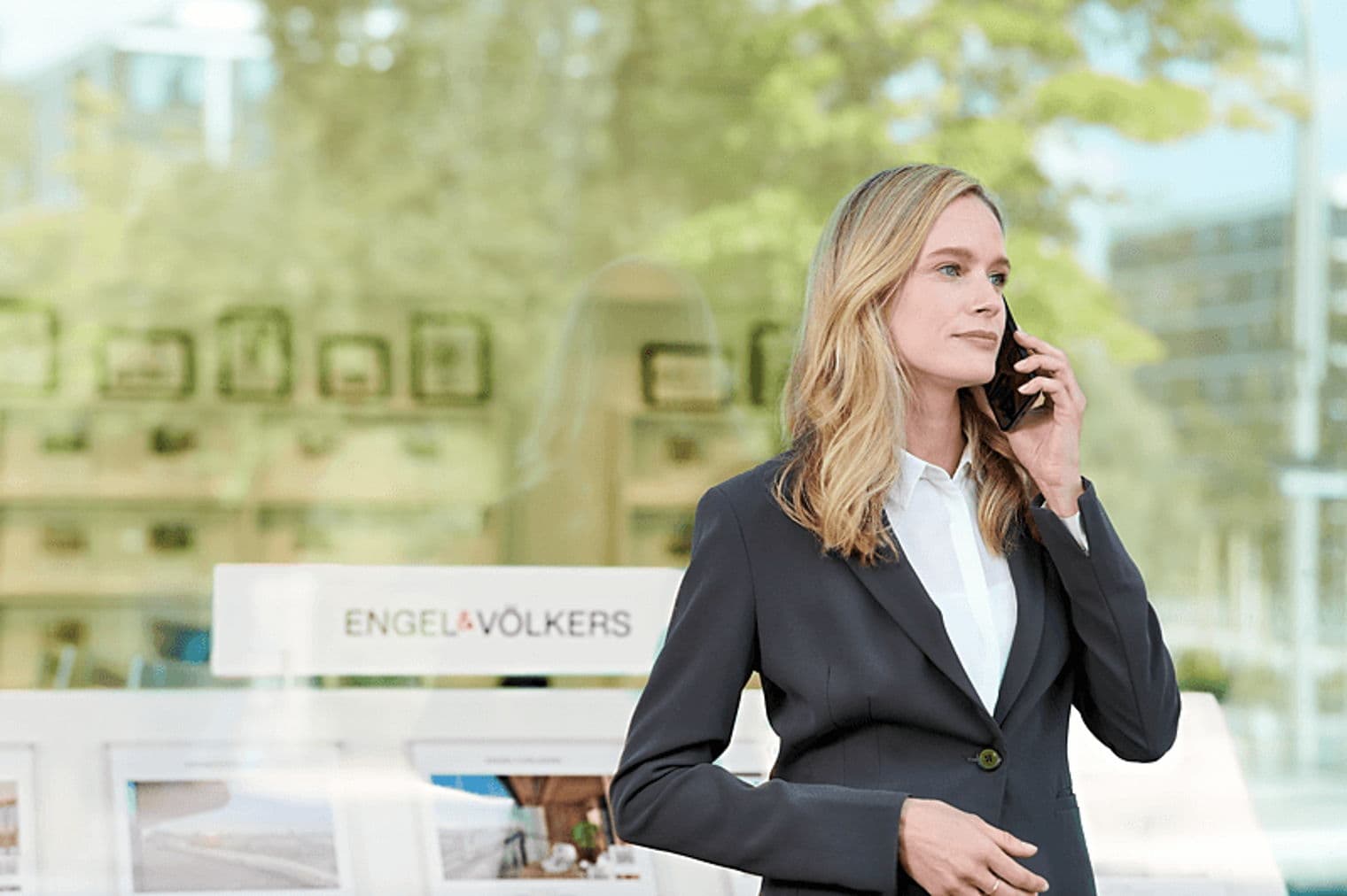 A blonde woman in a suit speaks on her phone in front of an Engel & Völkers real estate office.