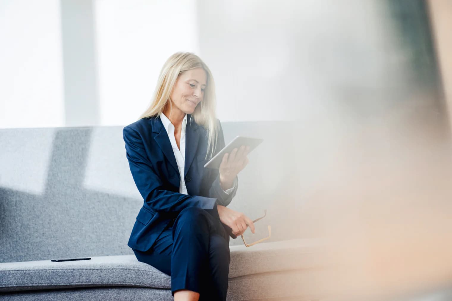 A blonde woman in a blue suit sits on a gray couch, looking at a tablet. She holds glasses in her hand.