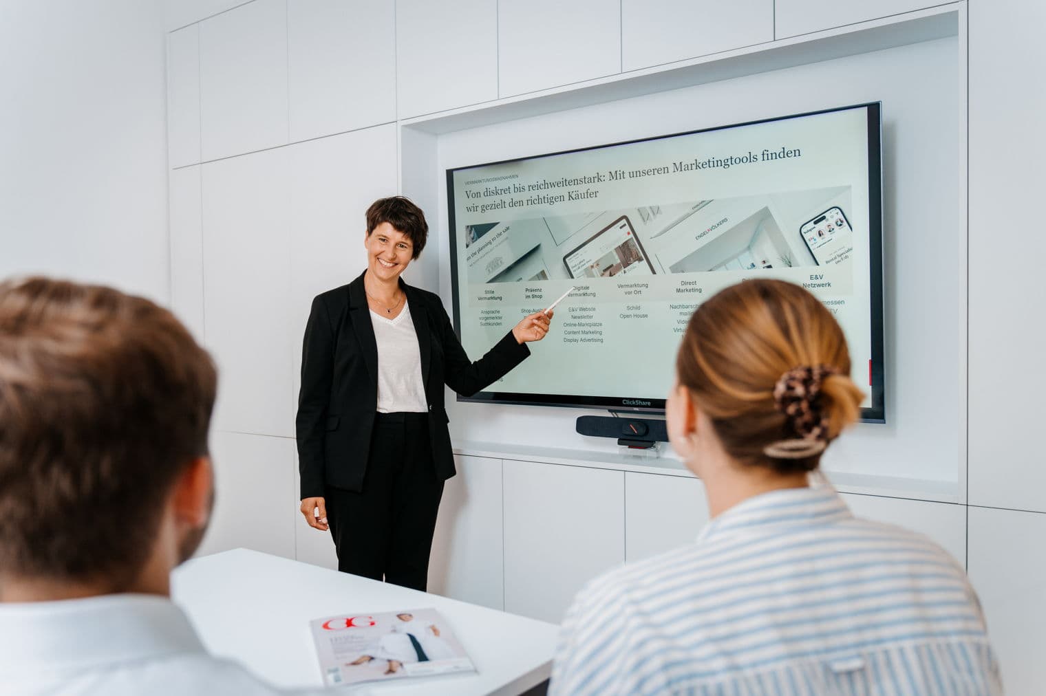 Eine Frau in einem schwarzen Anzug präsentiert zwei Personen Marketing-Tools auf einem großen Bildschirm.