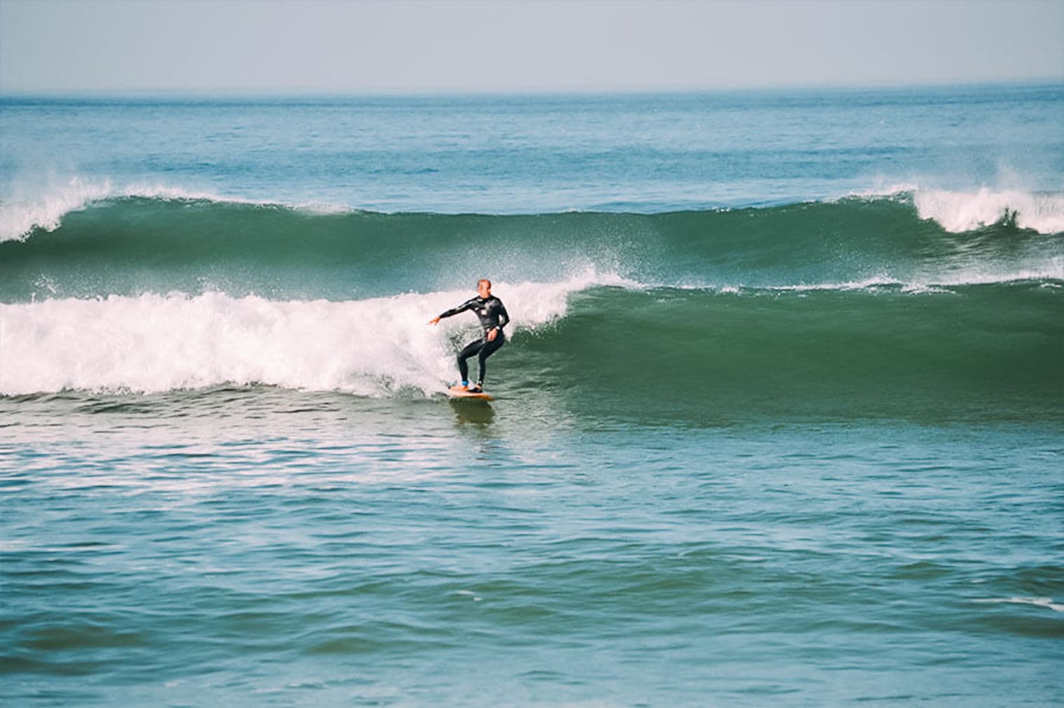 A surfer in a black wetsuit rides a wave on a light brown surfboard in the ocean.