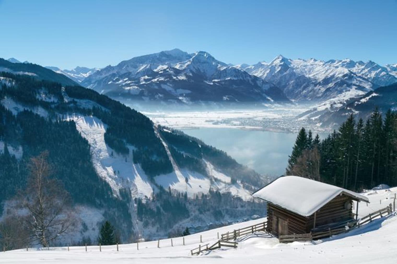 Schneebedeckte Berglandschaft mit einer Holzhütte im Vordergrund. Ein See und eine Stadt sind im Tal darunter sichtbar.