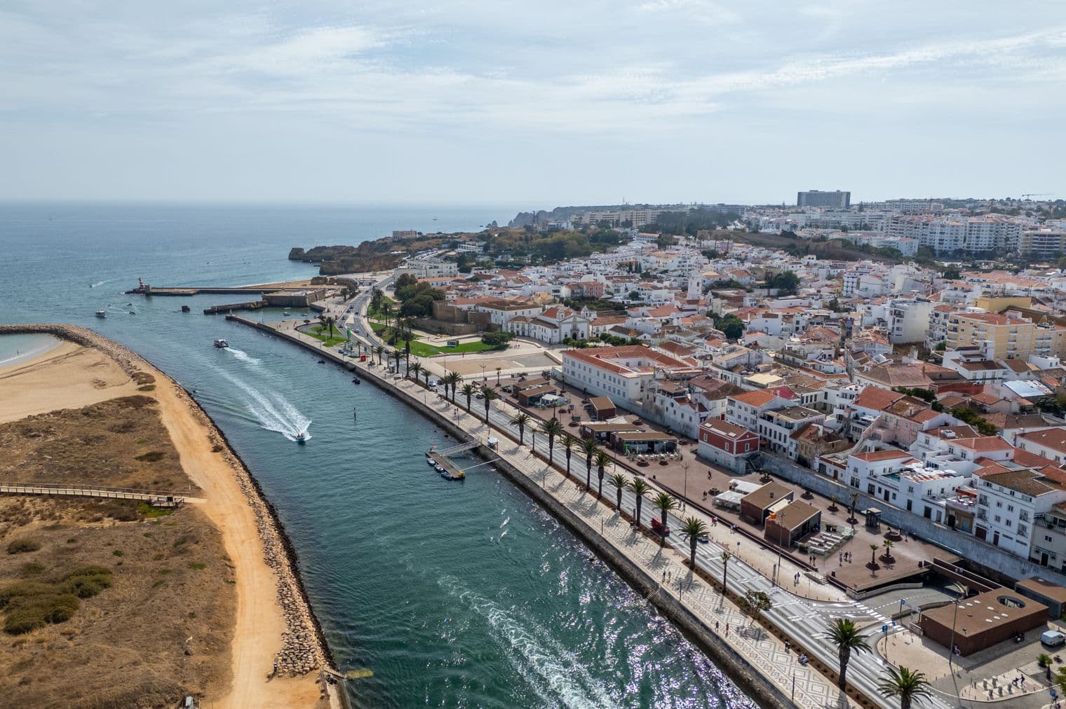 Aerial view of Lagos, Portugal, featuring a river, palm-lined promenade, and buildings with red-tiled roofs under a partly cloudy sky.