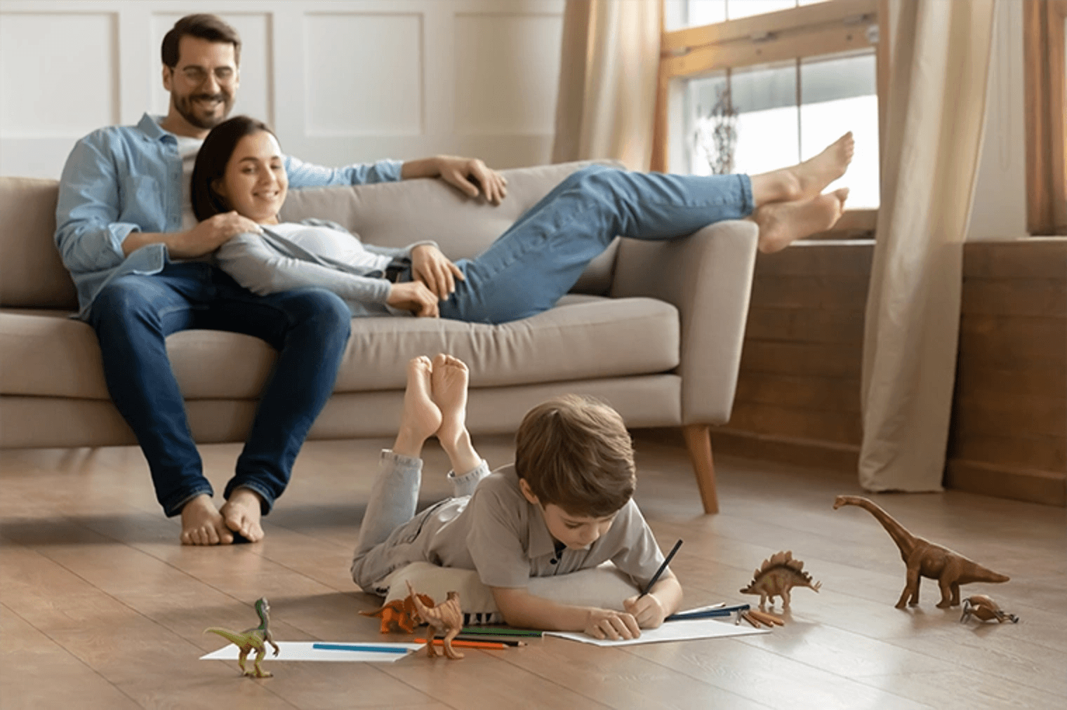 Eine Familie entspannt sich in ihrem Wohnzimmer. Ein Paar sitzt auf einem beigen Sofa, während ihr Sohn mit Dinosaurierspielzeug auf dem Boden malt.