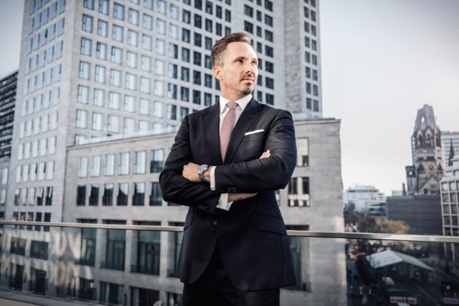 Ein Mann im Anzug steht mit verschränkten Armen auf einem Balkon, mit Stadtgebäuden im Hintergrund.