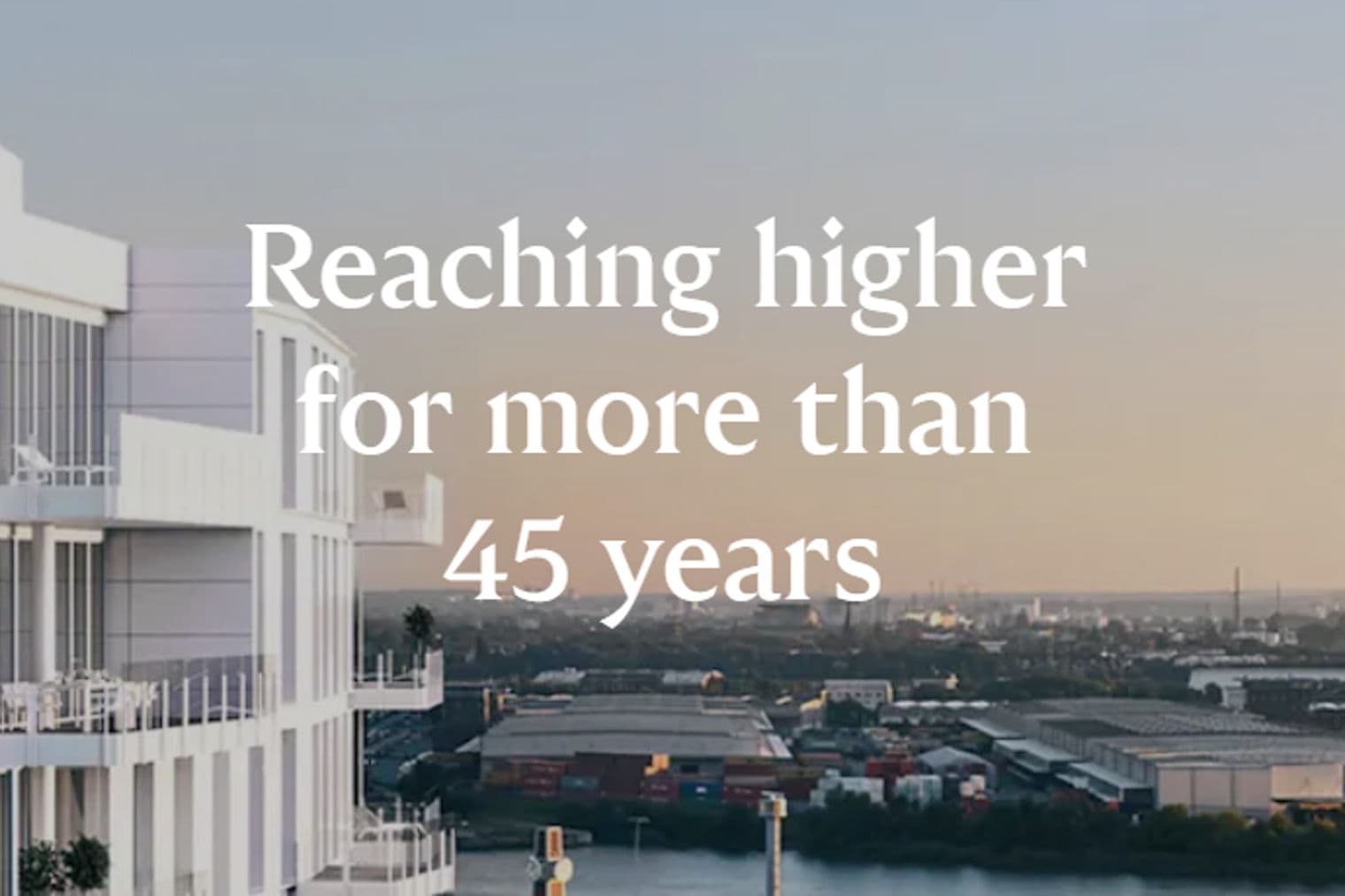 Edifício branco moderno com varandas com vista para o horizonte da cidade e o rio ao pôr do sol. A sobreposição de texto diz: "Alcançando mais alto por mais de 45 anos."