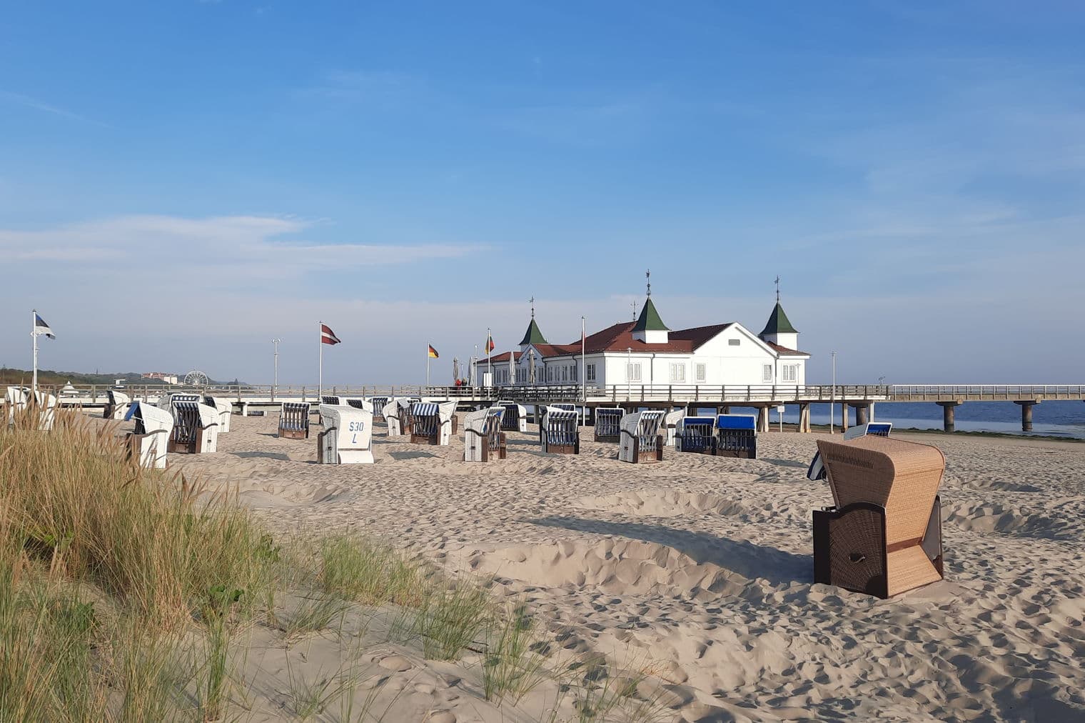 Sandy beach with many beach chairs, leading to a white pier building with flags under a blue sky.