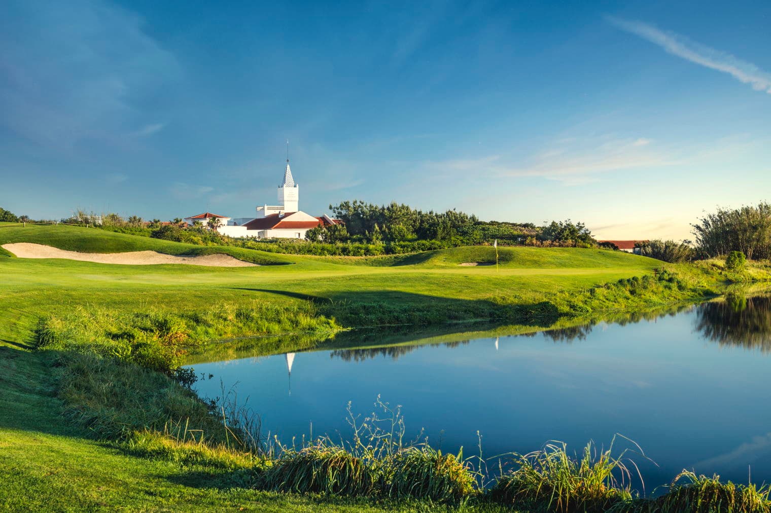 Golf course view with a white building and spire reflected in a pond under a blue sky.