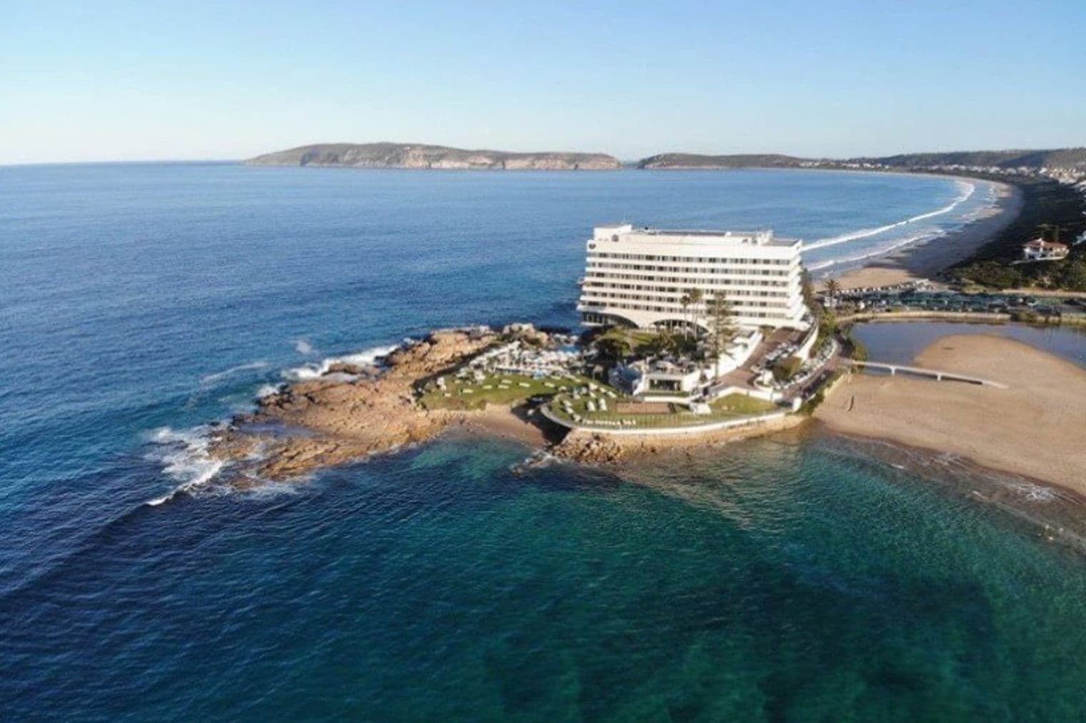 Aerial view of a white hotel on a rocky peninsula, turquoise ocean, sandy beach, and blue sky.