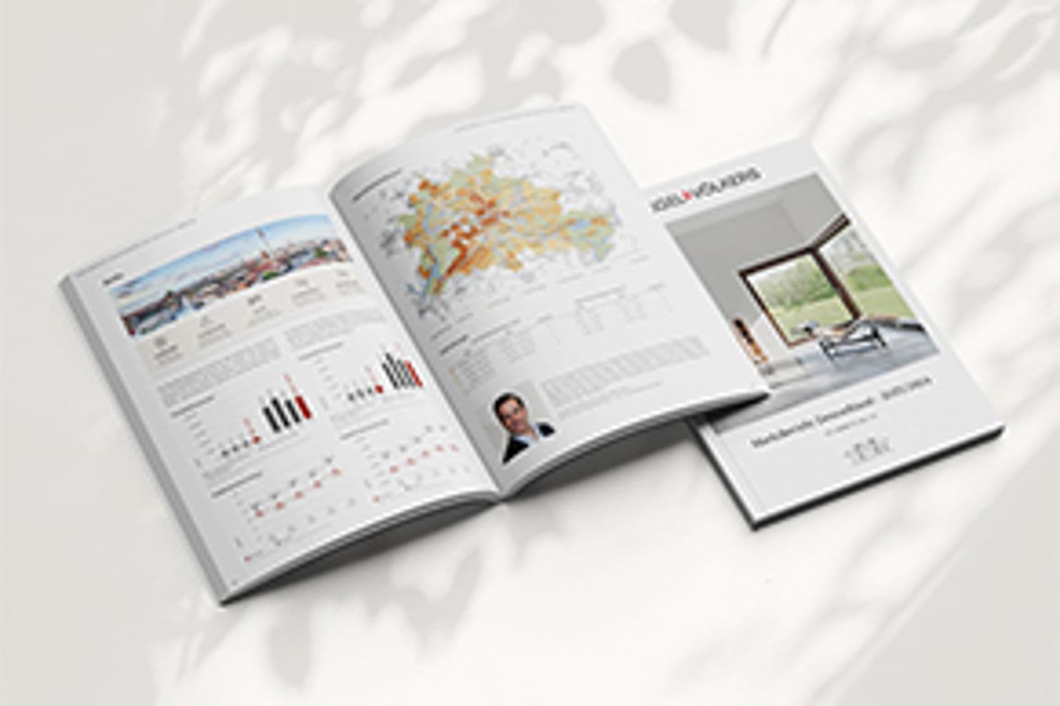 Geöffnete Immobilienbroschüre mit Diagrammen, Karten und Immobilienfotos, neben einer geschlossenen Broschüre auf einer weißen Oberfläche mit Blätterschatten.