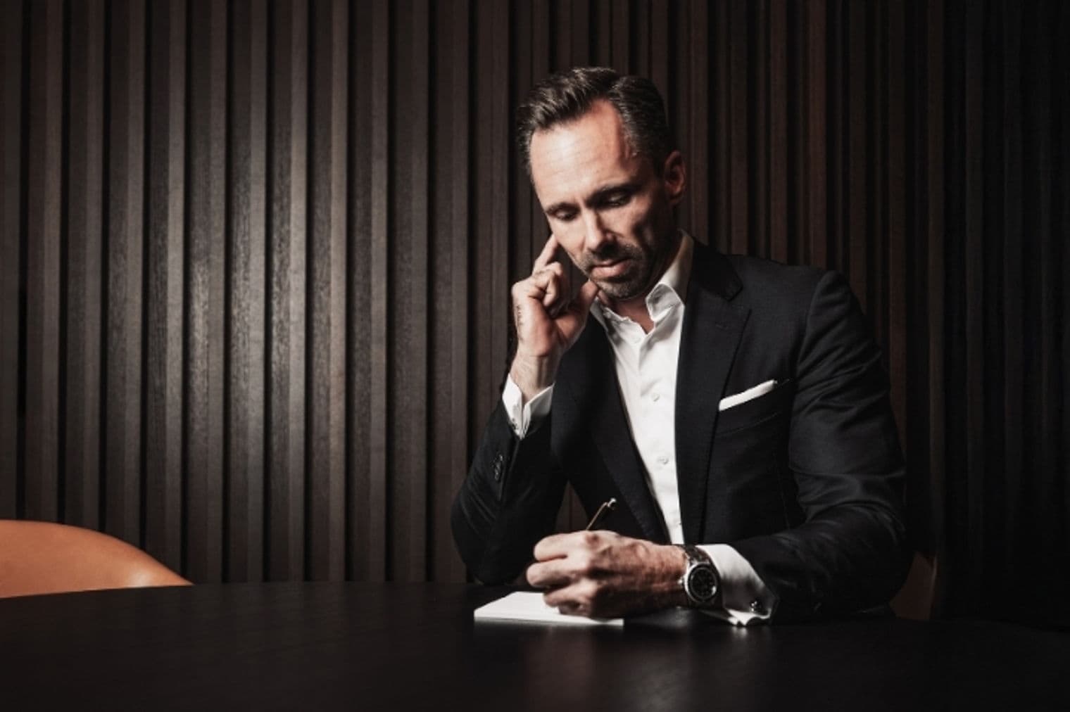 A man in a suit sits at a table, pen in hand, thinking and writing on a notepad.