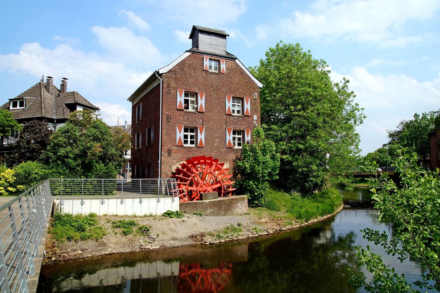 Backsteingebäude mit einem roten Wasserrad an einem Flussufer. Weiß umrandete Fenster und rote Fensterläden. Bäume und eine Brücke sind sichtbar.