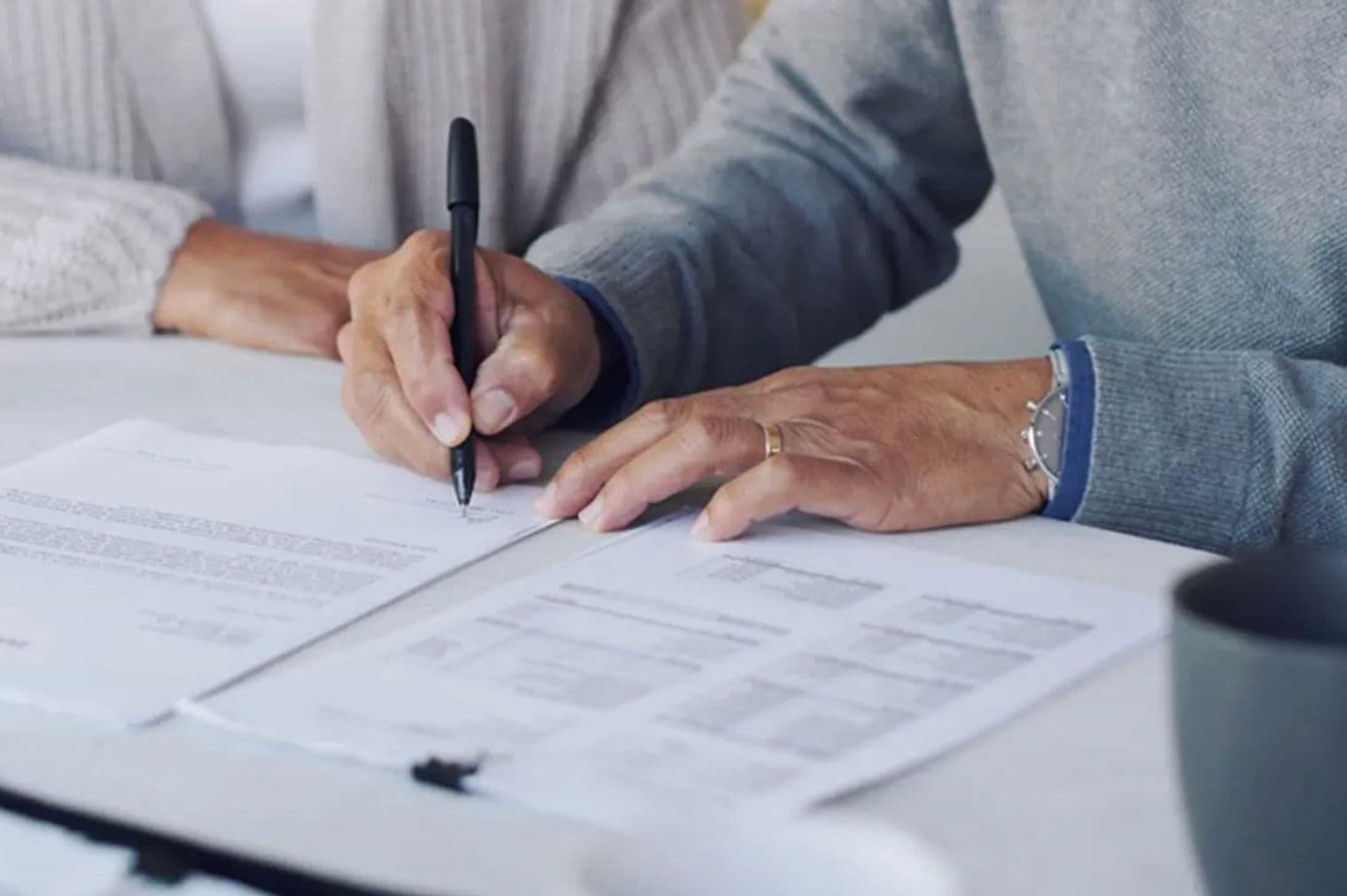 Gros plan sur un couple mature signant des documents avec un stylo noir sur une table blanche.