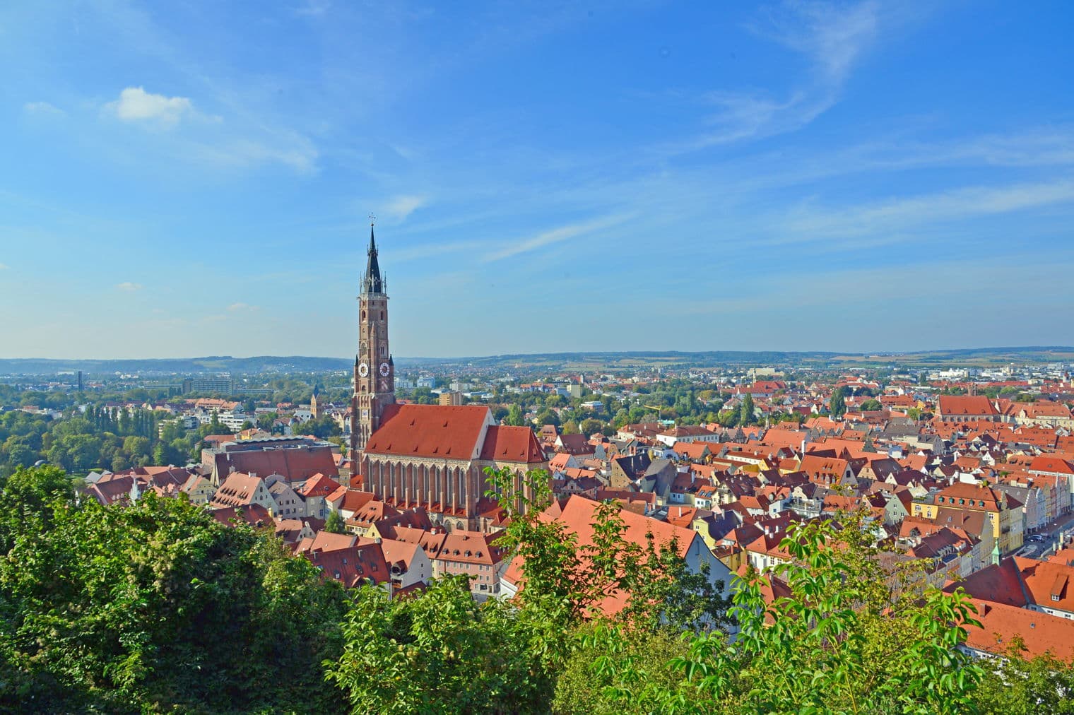 Ein Panoramablick auf Landshut, Deutschland, mit rot gedeckten Gebäuden, einem hohen Kirchturm und einem blauen Himmel.