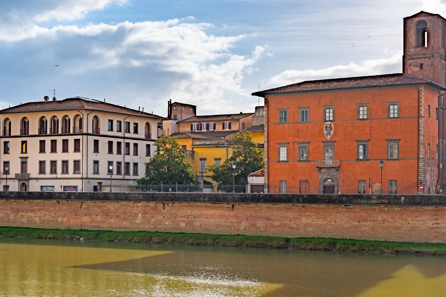 Flussblick auf farbenfrohe Gebäude in Pisa, Italien, unter einem bewölkten Himmel. Eine Ziegelmauer säumt das Flussufer.