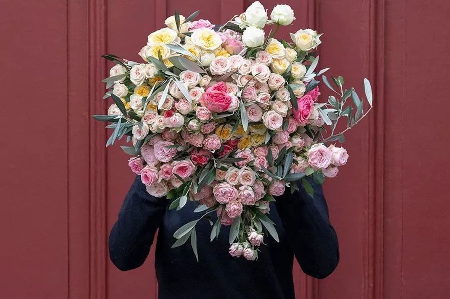 Eine Person mit schwarzem Pullover hält einen großen Blumenstrauß aus rosa, gelben und weißen Rosen vor einer rot getäfelten Tür.