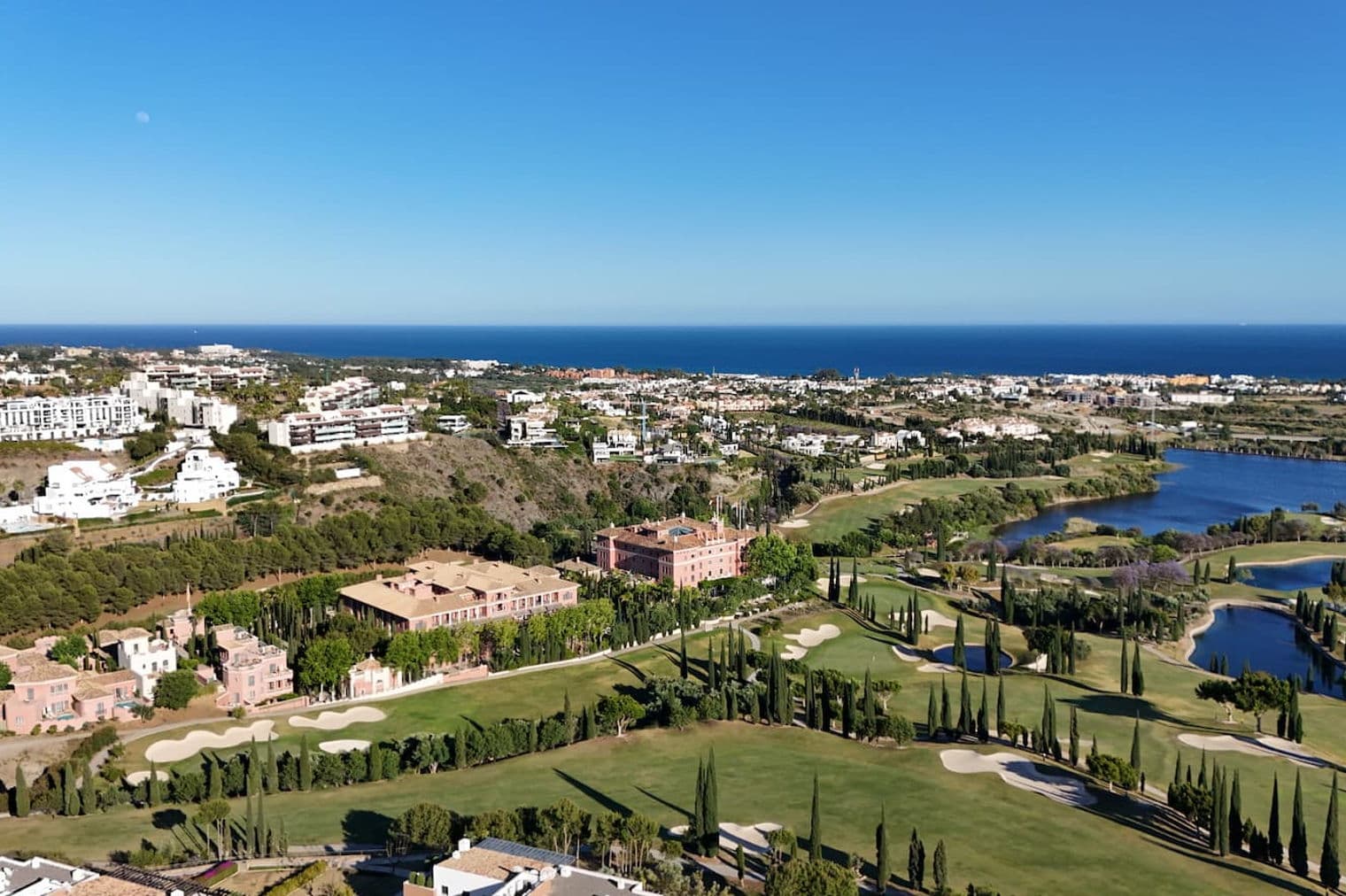Vista aèria d'un complex turístic de luxe amb un camp de golf, un llac i l'oceà al fons sota un cel blau clar.