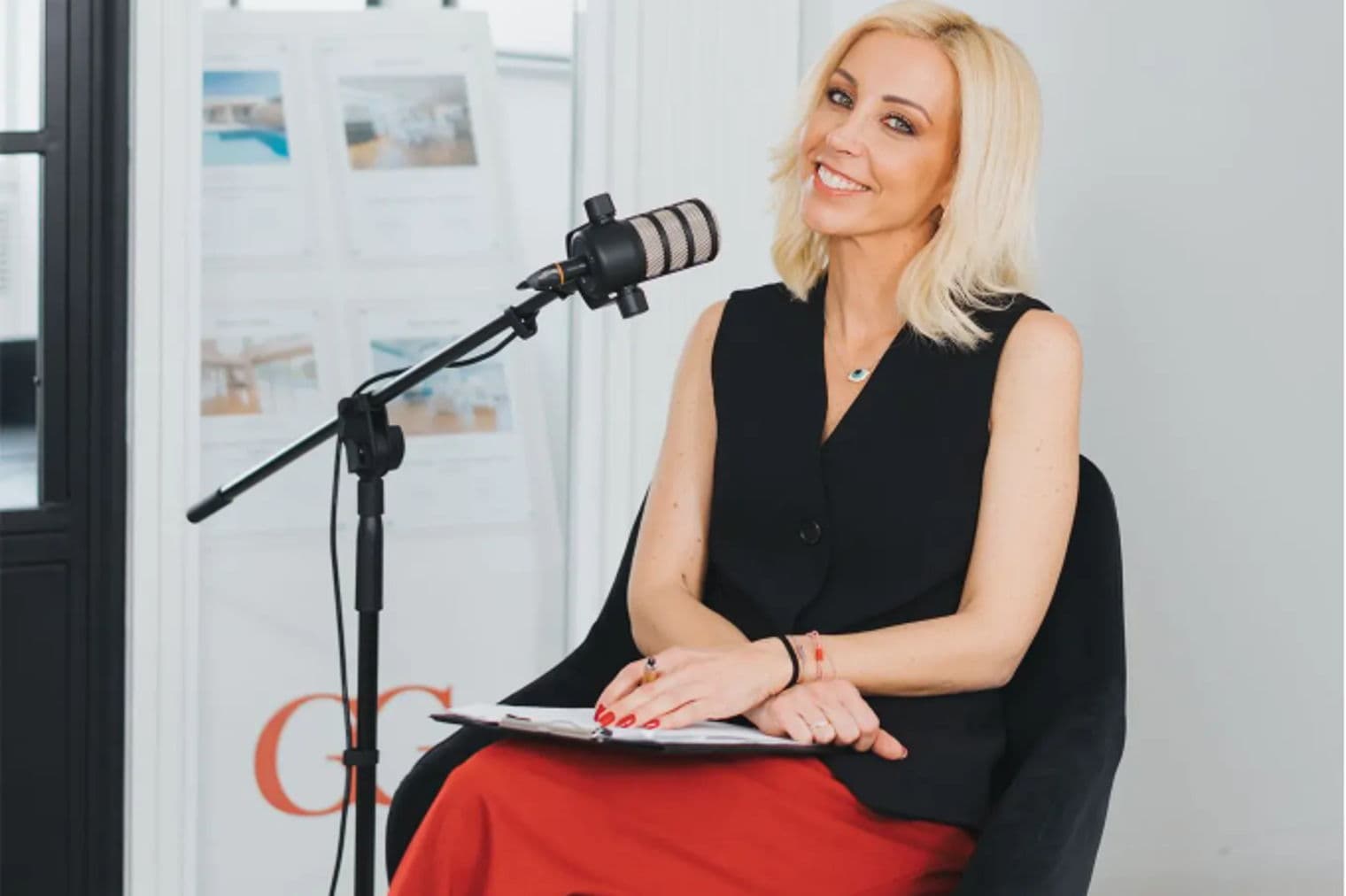Μια ξανθιά γυναίκα με μαύρο γιλέκο και κόκκινο παντελόνι κάθεται σε μια καρέκλα με ένα μικρόφωνο, χαμογελώντας. Φυλλάδια για ακίνητα βρίσκονται στο φόντο.