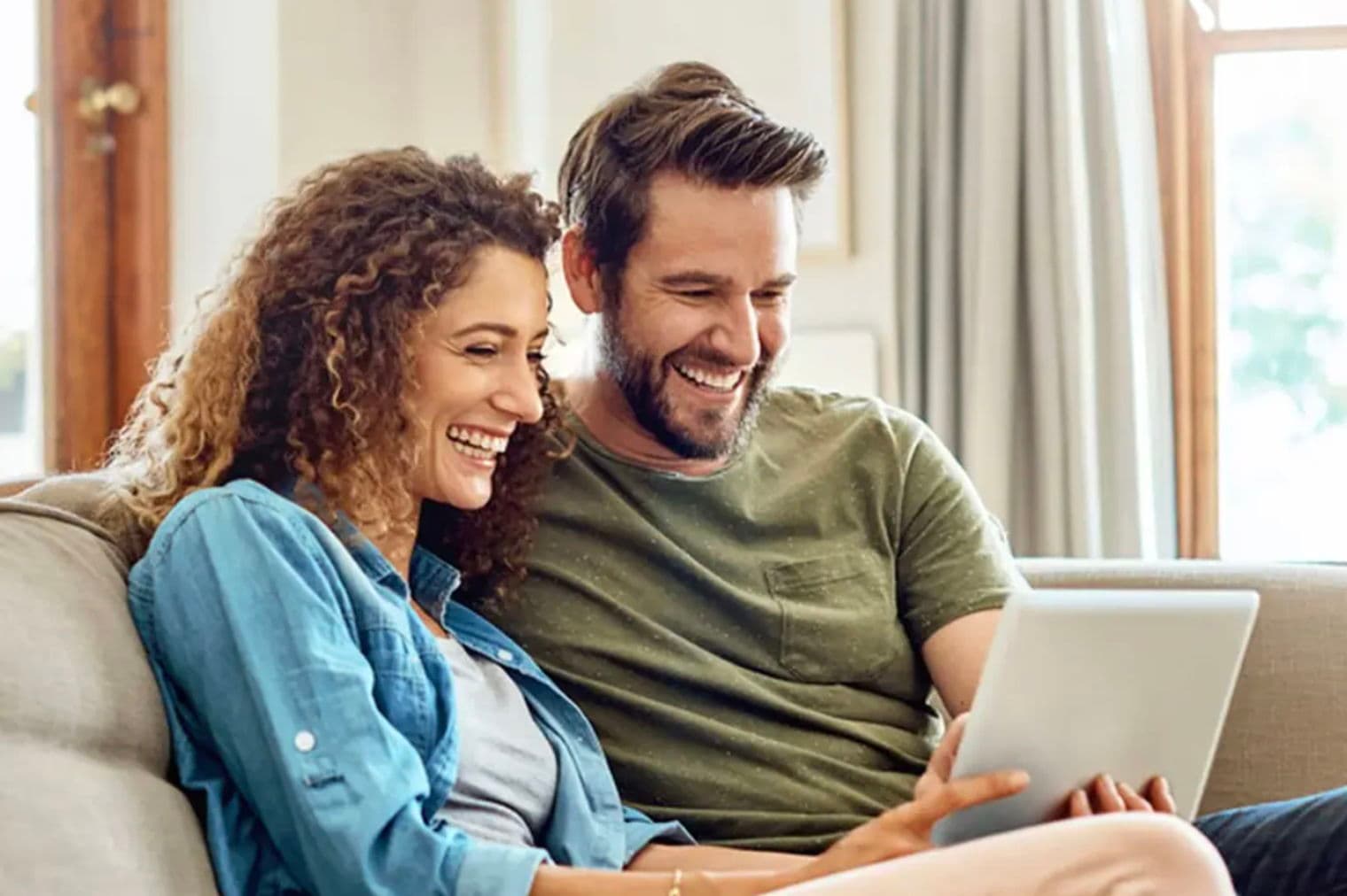 Un couple souriant est assis sur un canapé et regarde une tablette. La femme a les cheveux bouclés et porte une chemise bleue. L'homme a une barbe et porte une chemise verte.