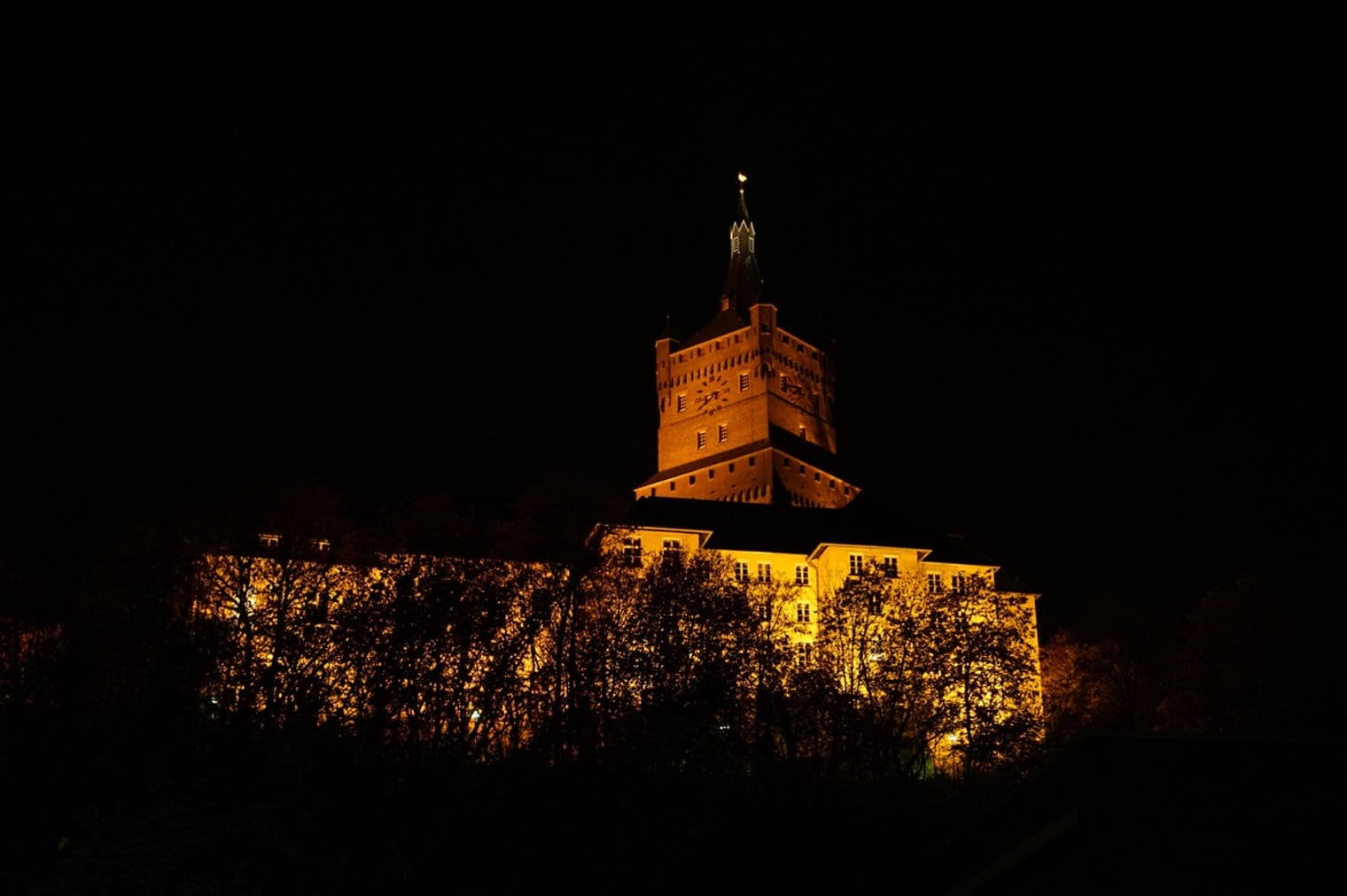 Nachtansicht einer beleuchteten mittelalterlichen Burg mit hohem Turm und Uhr, teilweise von Bäumen verdeckt.