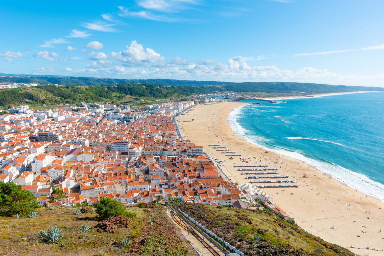 Aerial view of Nazaré, Portugal, showing red-roofed buildings, a sandy beach with people, and blue ocean under a partly cloudy sky.