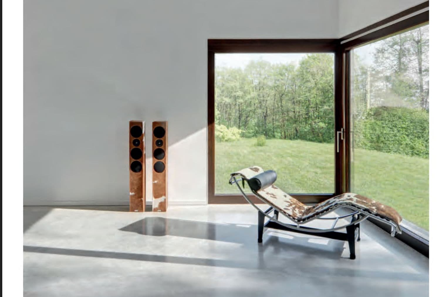 Minimalistischer Raum mit Lautsprechern, Chaiselongue und großen Fenstern mit Blick auf eine grüne Landschaft. Engel & Völkers Logo oben.