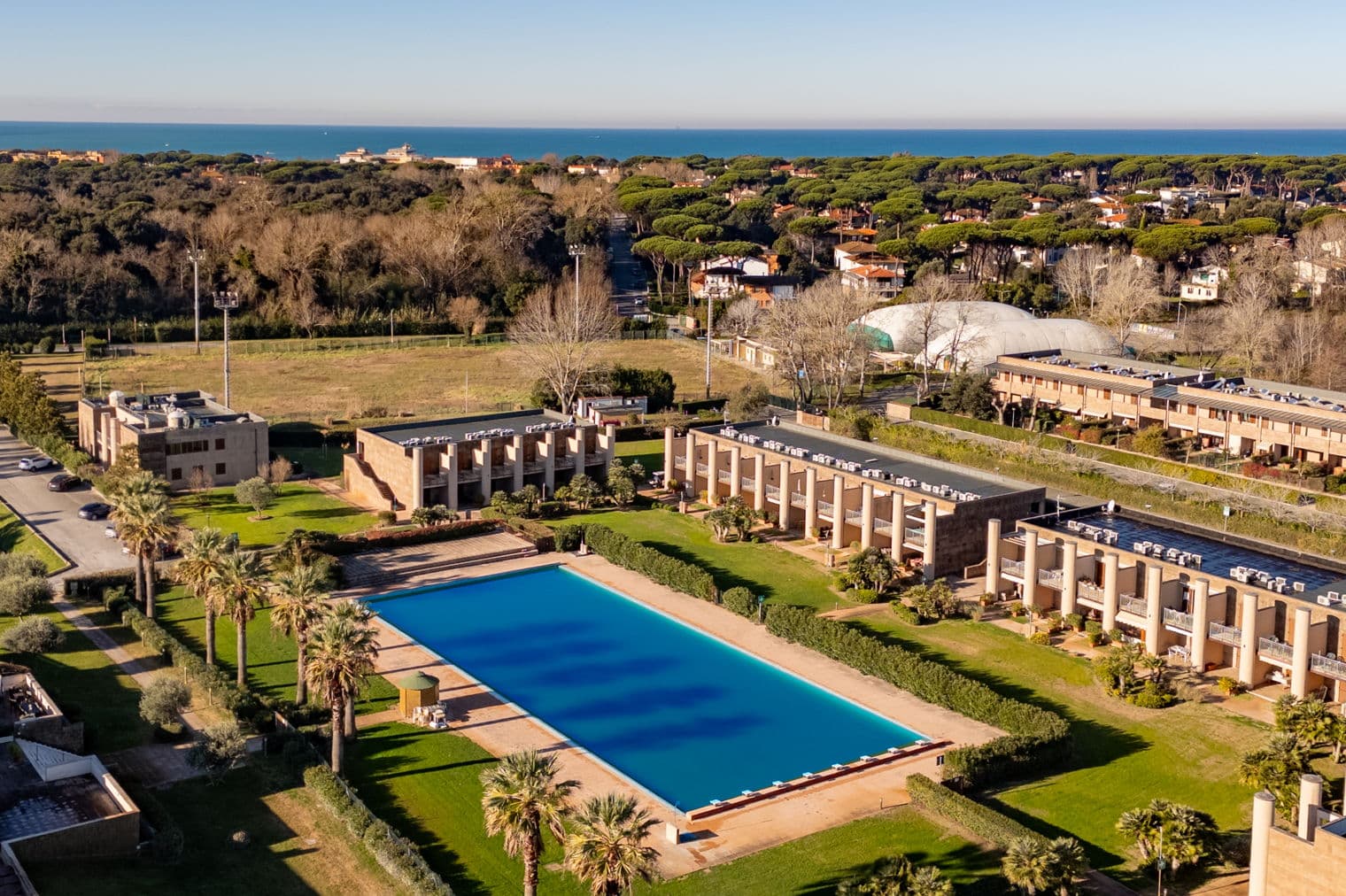 Luftaufnahme eines Resorts mit einem großen blauen Pool, Palmen und Gebäuden, mit dem Ozean im Hintergrund.