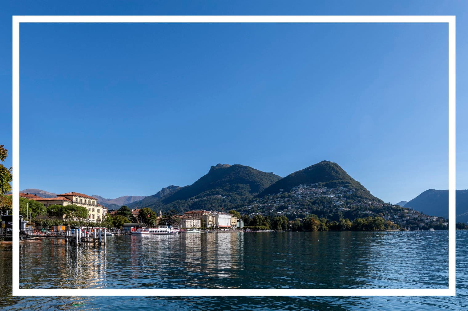 Blick auf den Luganersee mit Gebäuden, Bergen und einem klaren blauen Himmel. Das Wasser spiegelt die Gebäude und den Himmel wider.