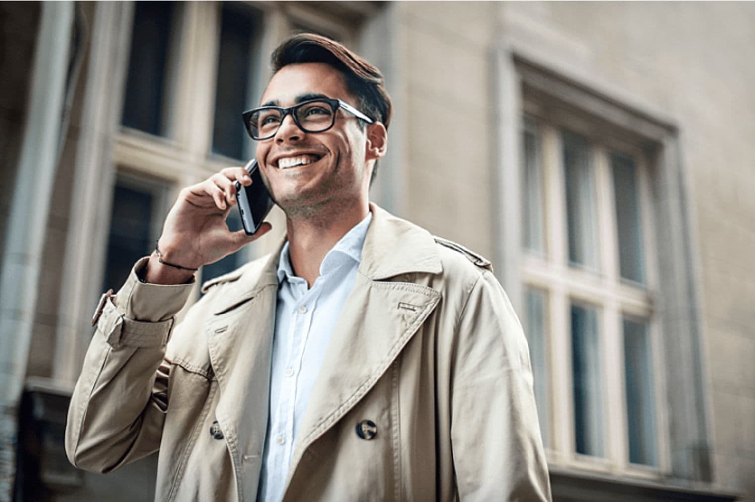 Lächelnder Mann mit Brille und Trenchcoat telefoniert vor einem Gebäude mit einem Handy.