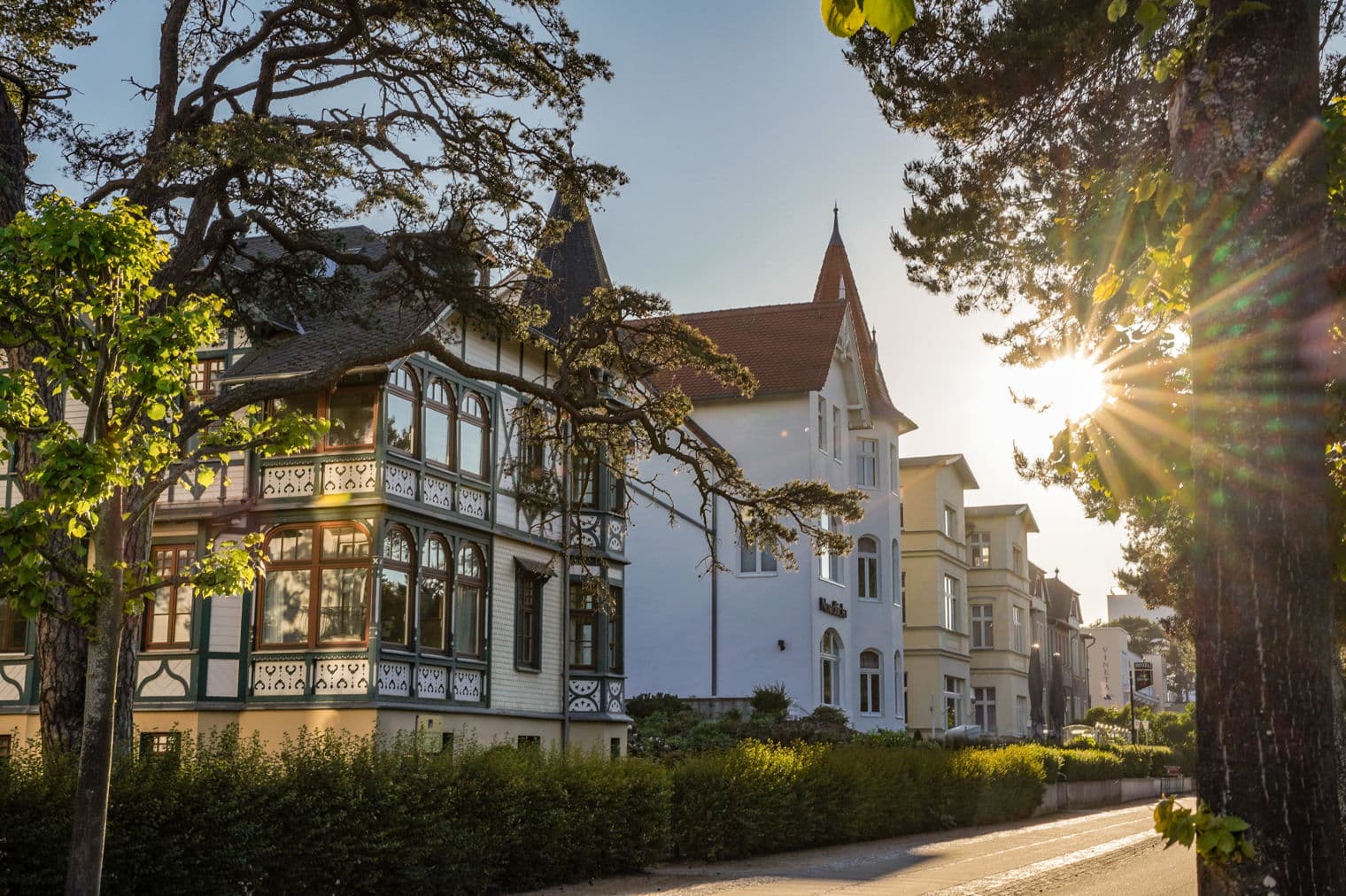 Straßenansicht von weißen Gebäuden mit roten Dächern, umrahmt von Bäumen und Sonnenlicht.
